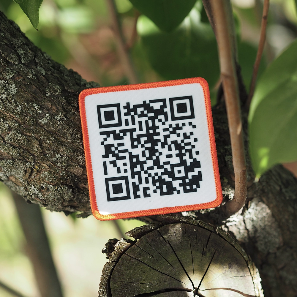 Painettu QR-koodi haalarimerkki, oranssit reunat, neliön muotoinen, taustalla puun oksia.