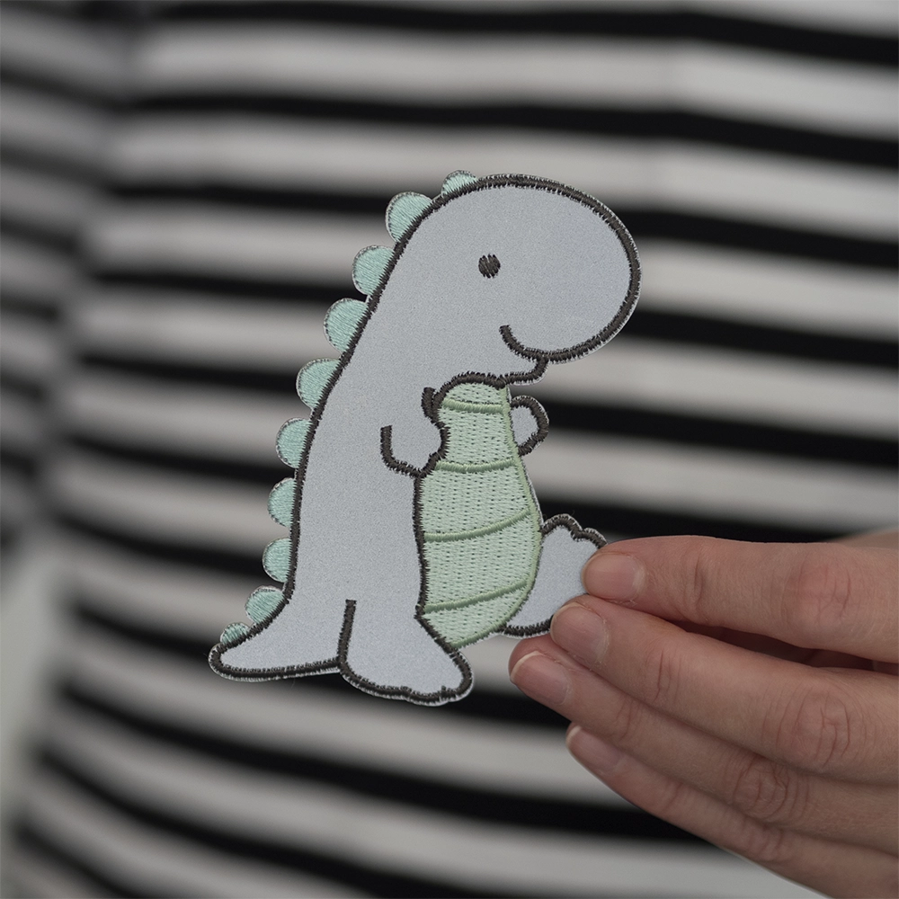 Heijastava brodeerattu dinosaurus haalarimerkki, vihreät yksityiskohdat. Merkki henkilön kädessä.