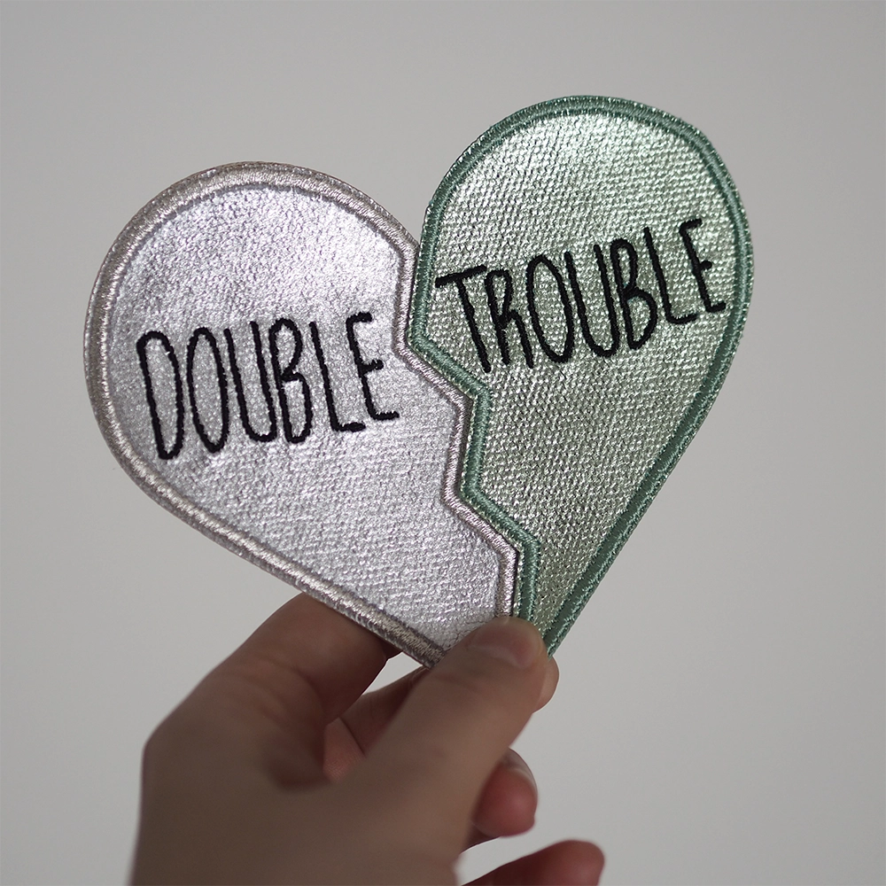 Metallinhohtoinen brodeerattu kaksiosainen haalarimerkki, merkeissä tekstit: Double ja Trouble. Merkit henkilön kädessä.