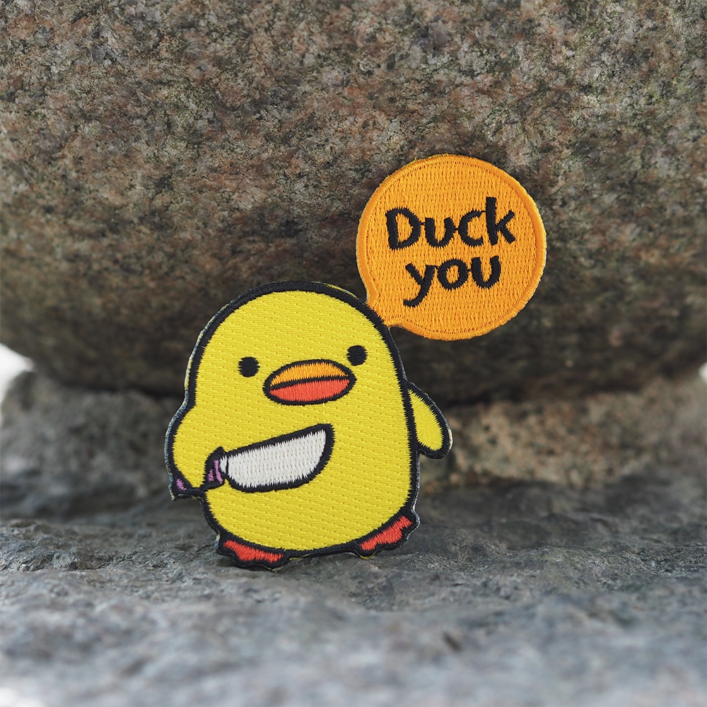 Brodeerattu tipu-aiheinen haalarimerkki, puhekuplassa teksti: Duck you. Taustalla kivi.