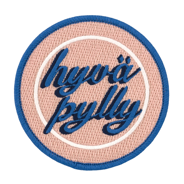 Brodeerattu pyöreä haalarimerkki, teksti: Hyvä pylly. Värimaailmassa sinistä, persikan sävyä ja valkoista.