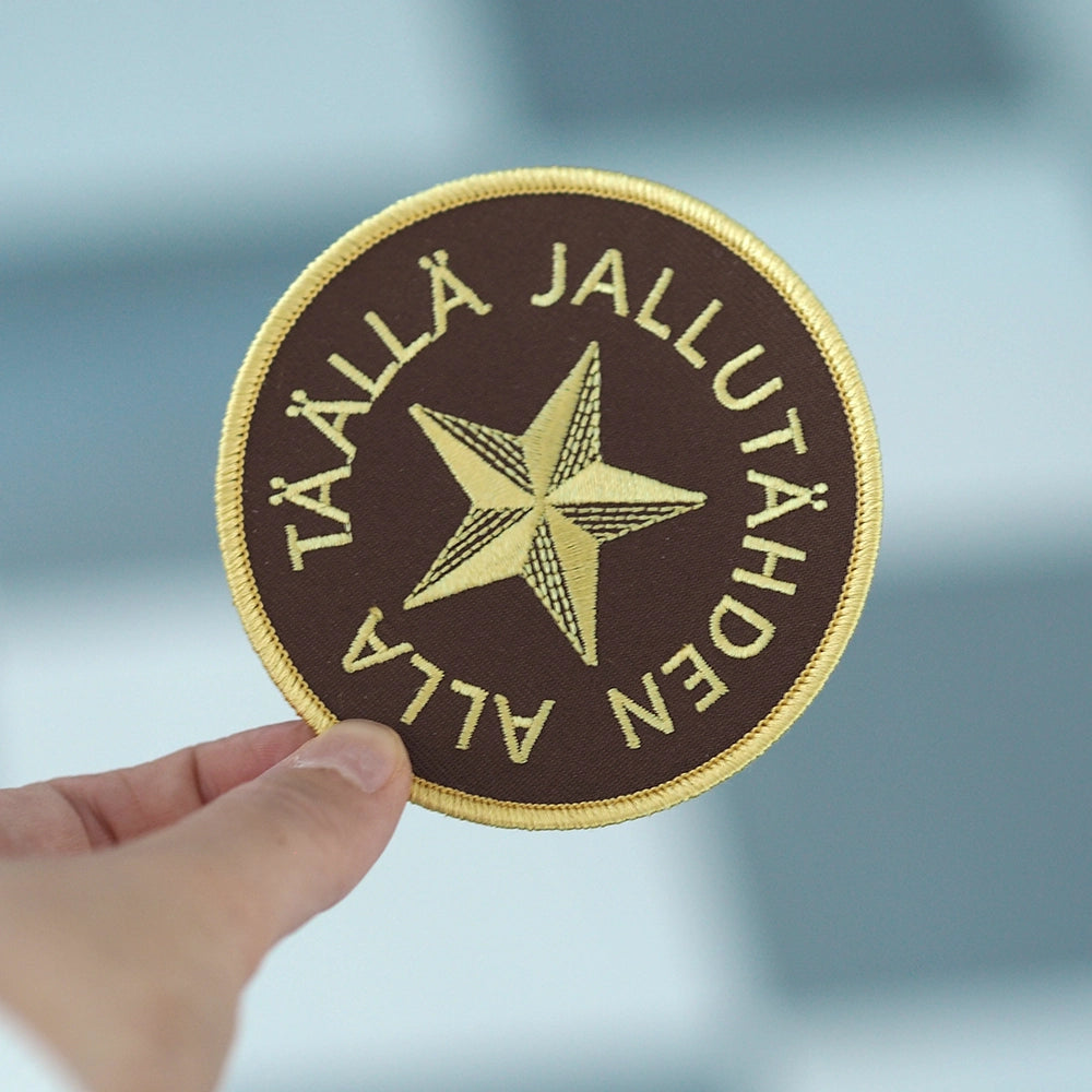 Brodeerattu haalarimerkki, jallutähti, teksti: Täällä jallutähden alla. Merkki henkilön kädessä.