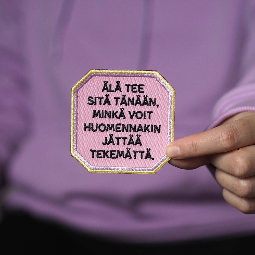 Brodeerattu haalarimerkki, teksti: Älä tee sitä tänään, minkä voit huomennakin jättää tekemättä. Merkki henkilön kädessä.