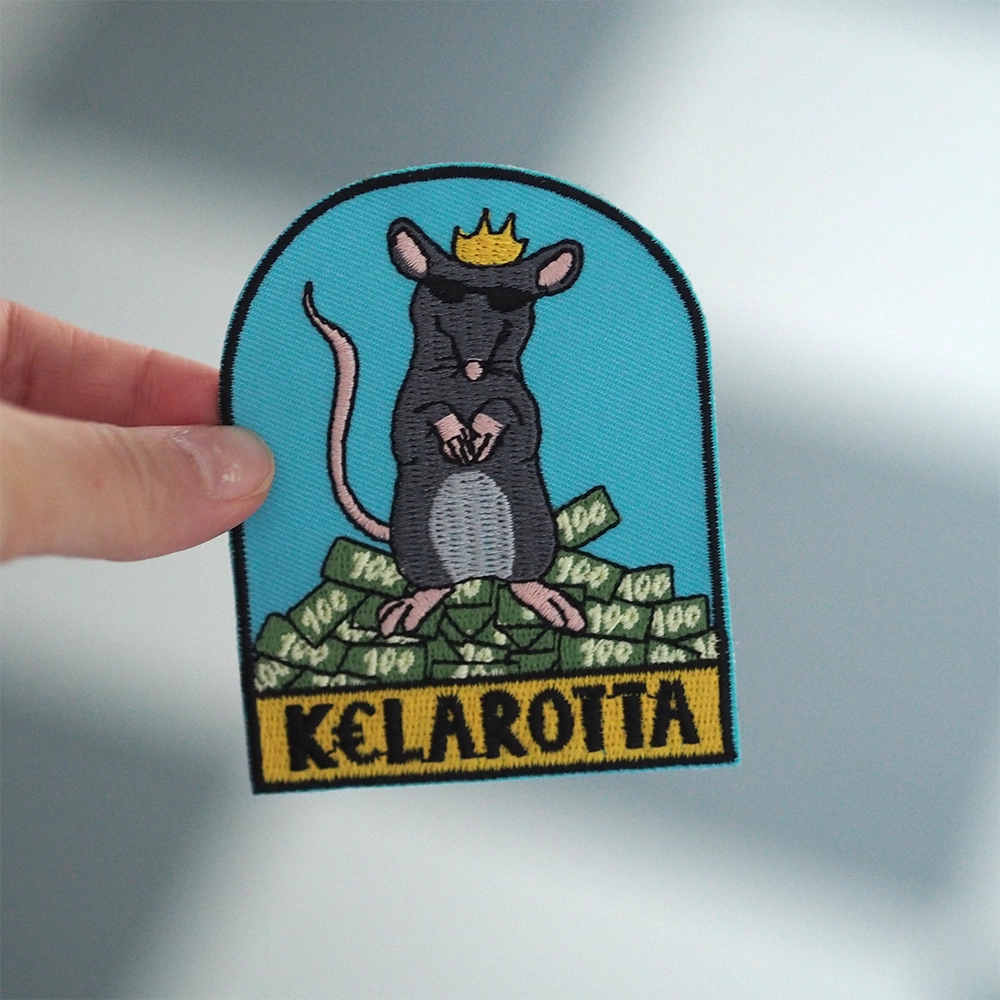 Brodeerattu rotta-haalarimerkki, kruunu, teksti: Kelarotta. Merkki henkilön kädessä.