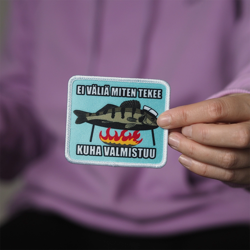 Painettu haalarimerkki, kuha nuotiolla, teksti: Ei väliä miten tekee kuha valmistuu. Merkki henkilön kädessä.