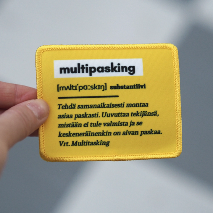 Painettu haalarimerkki, selitys siitä mitä on multipasking, keltainen. Merkki henkilön kädessä.