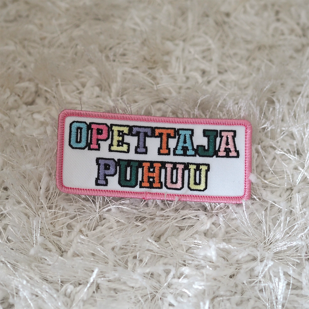 Brodeerattu haalarimerkki, teksti: Opettaja puhuu. Merkki maton päällä.