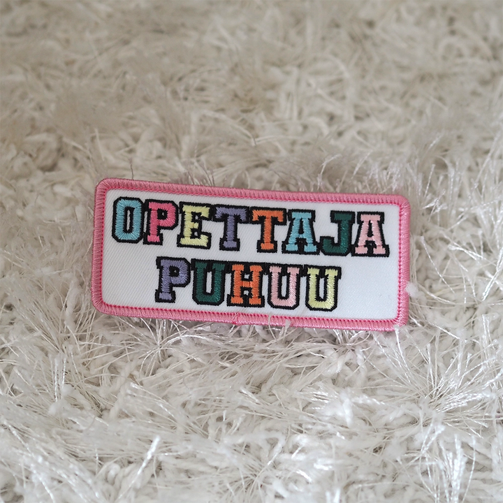 Brodeerattu haalarimerkki, teksti: Opettaja puhuu. Merkki maton päällä.