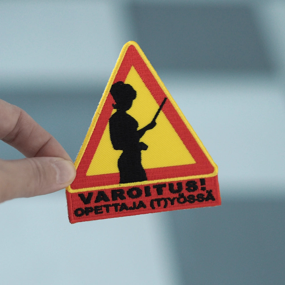 Brodeerattu haalarimerkki, varoituskolmio, teksti: Varoitus, opettaja (t)yössä. Merkki henkilön kädessä.