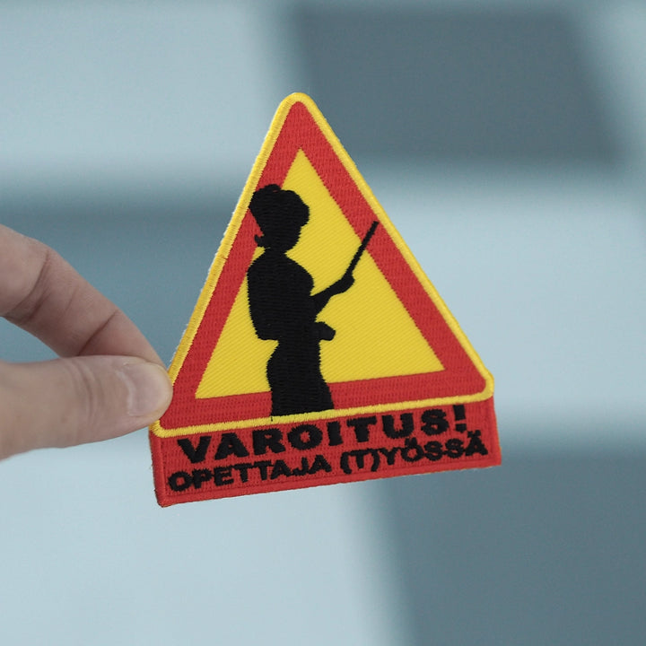 Brodeerattu haalarimerkki, varoituskolmio, teksti: Varoitus, opettaja (t)yössä. Merkki henkilön kädessä.