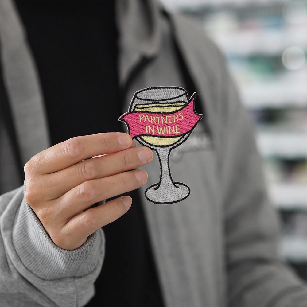 Brodeerattu viini-aiheinen haalarimerkki, viinilasi, teksti: Partners in wine. Merkki henkilön kädessä.