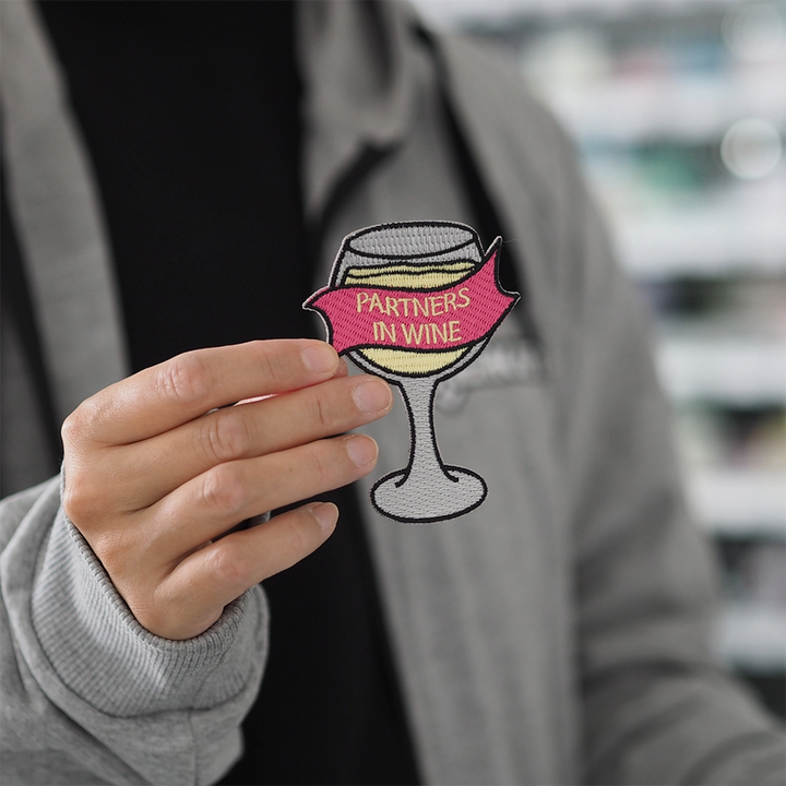 Brodeerattu viini-aiheinen haalarimerkki, viinilasi, teksti: Partners in wine. Merkki henkilön kädessä.