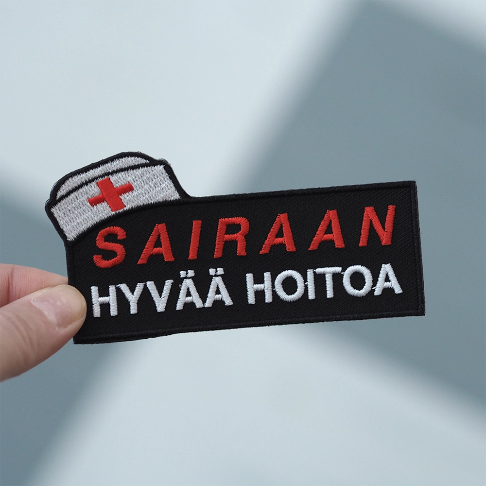 Brodeerattu haalarimerkki, teksti: Sairaan hyvää hoitoa. Merkki henkilön kädessä.