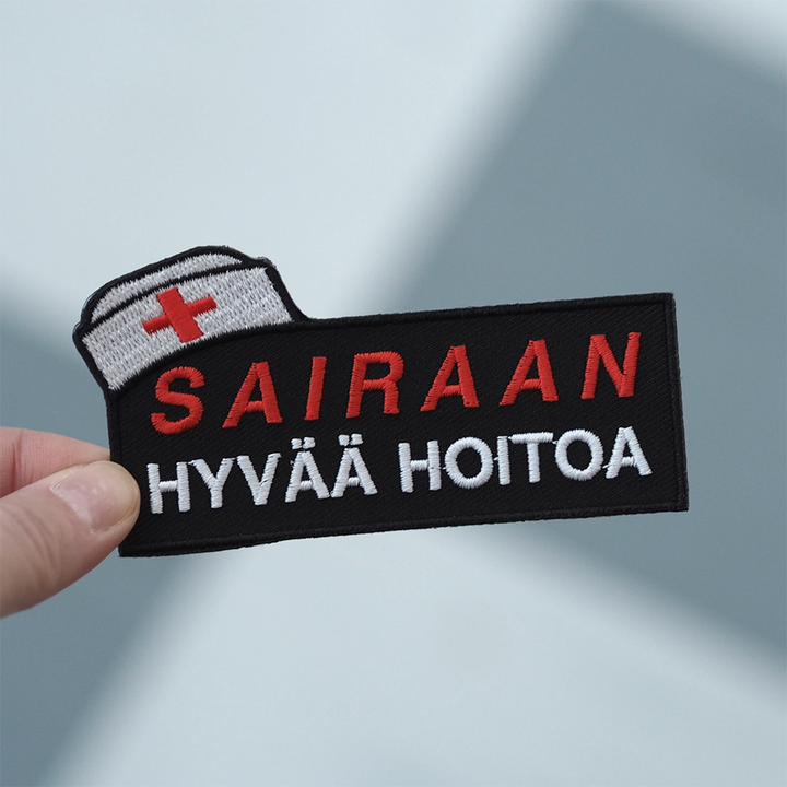 Brodeerattu haalarimerkki, teksti: Sairaan hyvää hoitoa. Merkki henkilön kädessä.