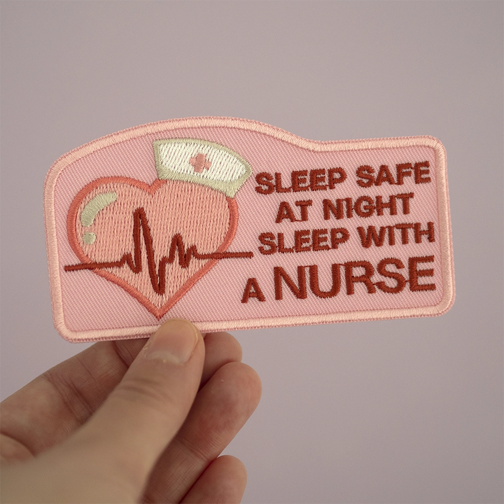 Brodeerattu sairaanhoitaja-aiheinen haalarimerkki, teksti: Sleep safe at night sleep with a nurse. Merkki henkilön kädessä.