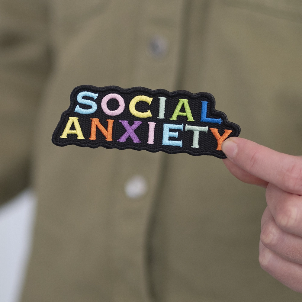 Brodeerattu haalarimerkki, teksti: Social anxiety. Merkki henkilön kädessä.