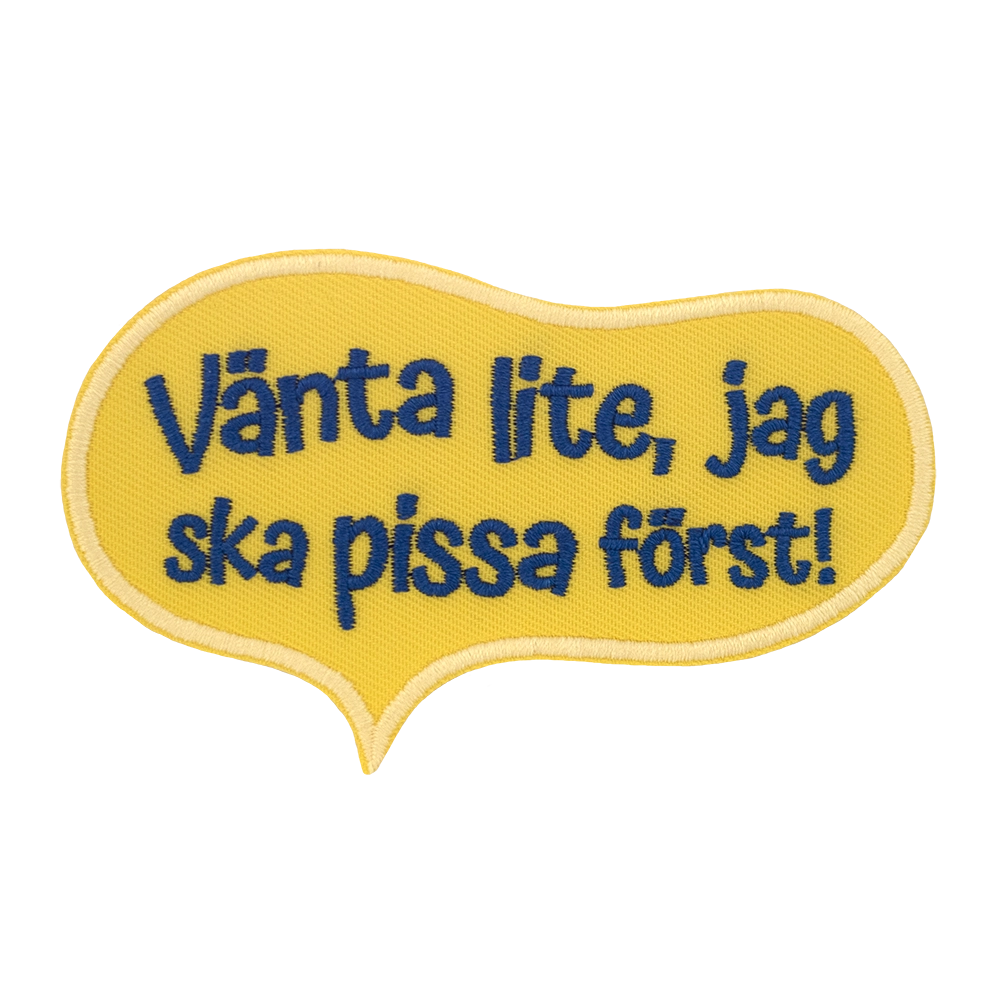 Brodeerattu keltainen haalarimerkki, teksti: Vänta lite, jag ska pissa först!