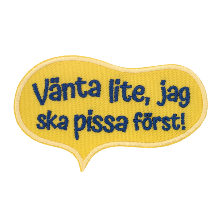 Brodeerattu keltainen haalarimerkki, teksti: Vänta lite, jag ska pissa först!