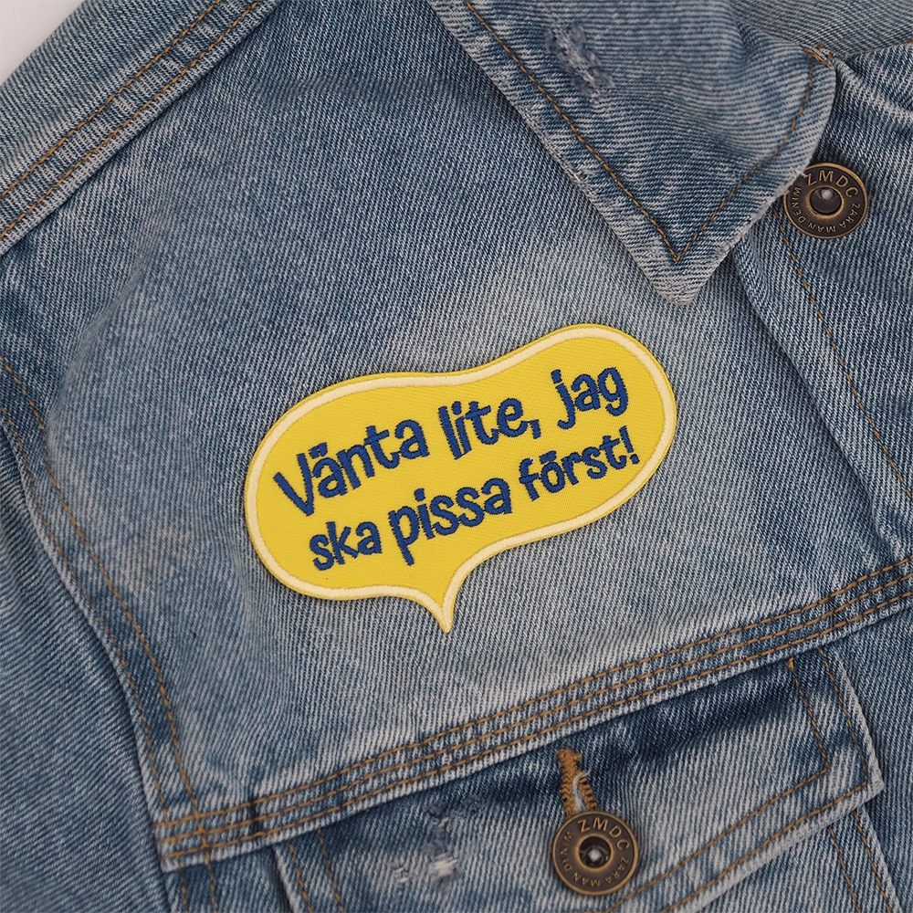 Brodeerattu keltainen haalarimerkki, teksti: Vänta lite, jag ska pissa först! Taustalla farkkutakki.